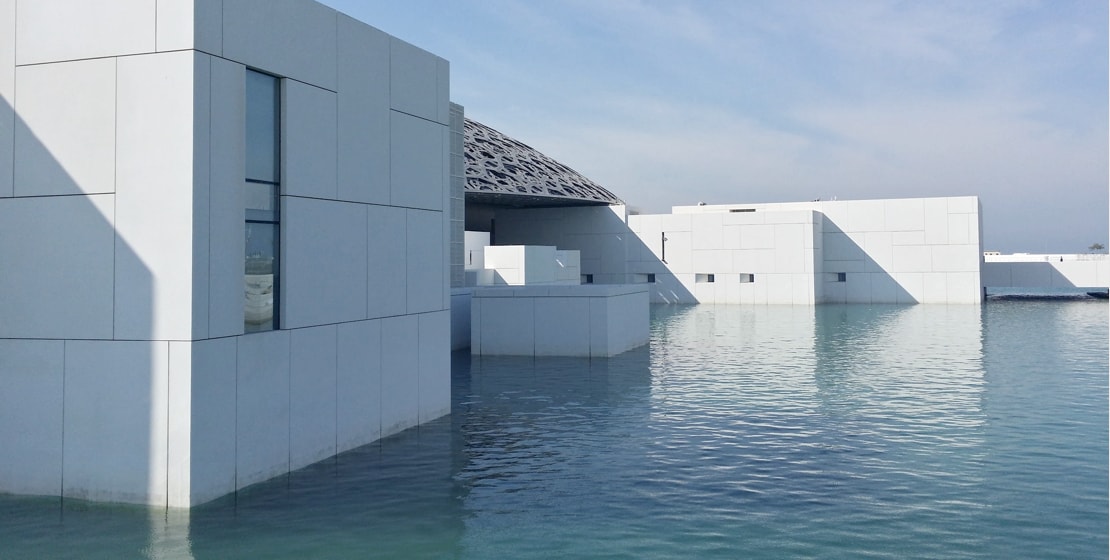 The image depicts modern, minimalist white buildings partially submerged in water, with a clear, blue sky in the background. The architecture has clean lines and flat surfaces, and the water reflects the buildings, creating a serene, mirror-like effect.