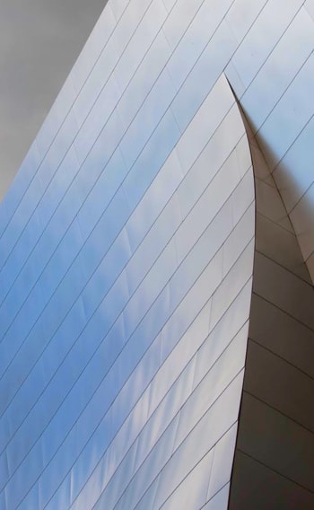 The image features a close-up view of a sleek, modern building with a smooth, curved surface. The structure appears to be made of metal, and the reflective surface captures the soft hues of the sky, creating a gradient of light and shadow.