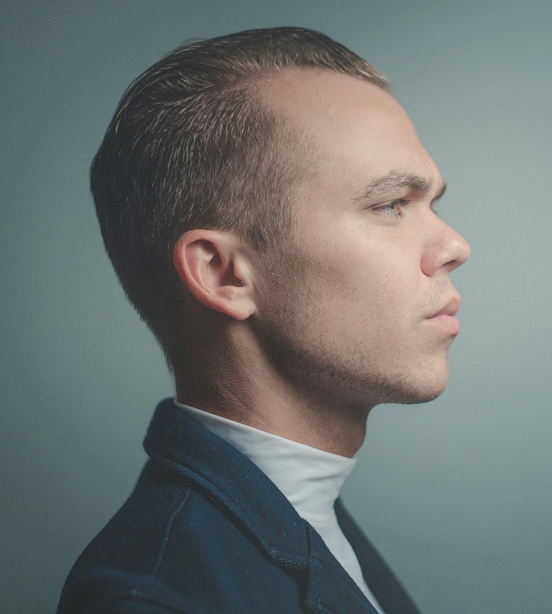 A profile photograph of the author with slicked-back hair and a contemplative expression, dressed in a dark jacket and a white high-neck shirt, against a muted gray background.